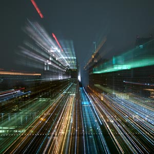Dieses Bild zeigt eine abstrakte Darstellung einer Stadt bei Nacht. Die Lichter der Gebäude und Straßen sind in langen Belichtungszeiten eingefangen, was zu leuchtenden, farbigen Streifen führt, die sich von der Mitte des Bildes nach außen bewegen. Die Farben umfassen Rot, Grün, Blau und Gelb, was eine dynamische und lebendige Atmosphäre erzeugt, die die Schnelligkeit und den Fortschritt der modernen Technologie symbolisieren könnte.